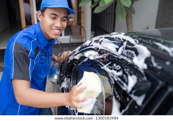Auto service staff in blue uniform\
cleaning car with sponge and foam at car owner\'s\
home