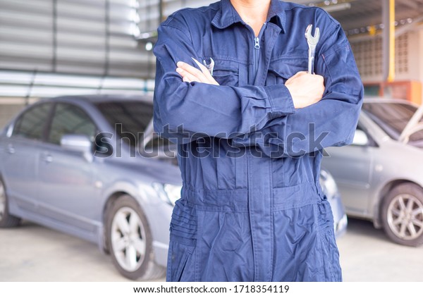 Auto service technician\
in uniform standing on the background of a car with a screwdriver\
and repair and maintenance of the car. Garage and car parts\
replacement concept
