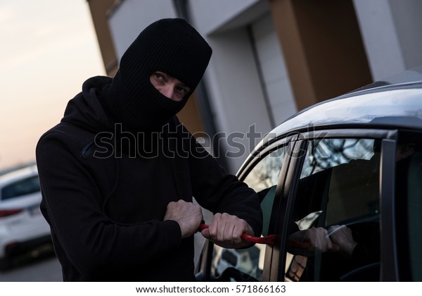 Auto thief in black balaclava trying to\
break into car with crowbar. Car thief, car\
theft
