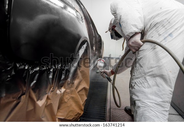 auto worker spraying black paint on a car\
in an auto garage with protective\
gear