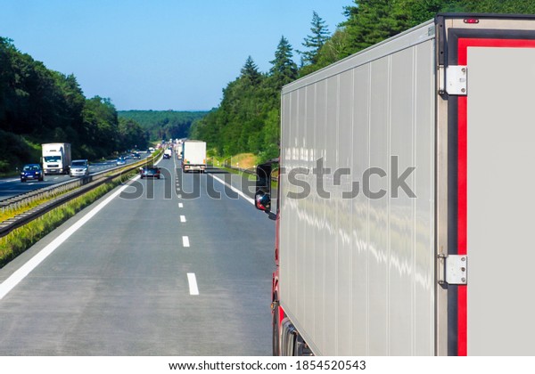 Autobahn in Europe. Cars and trucks, traffic on\
the highway. Truck on foreground, back view, asphalt road in rural\
forest landscape