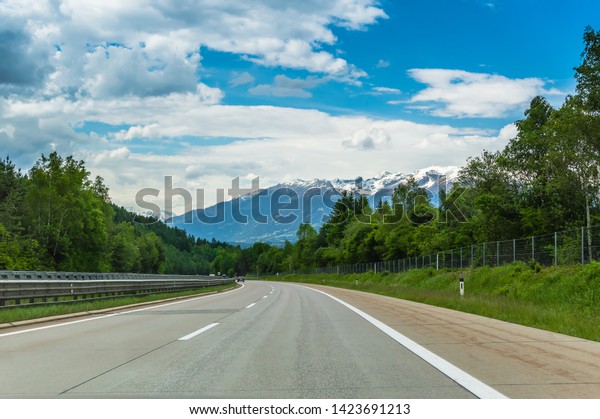 Autobahn or highway in the mountains with clear\
marking surrounded by vibrant green trees under blue sky. Stunning\
view and a snow-covered mountain in the background. The Alps,\
Austria