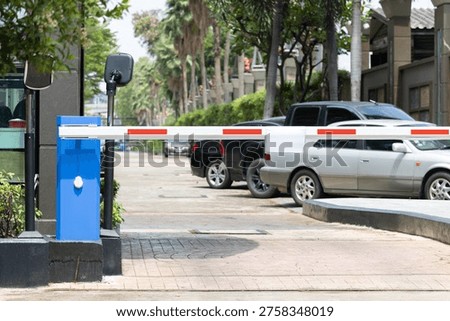 Automatic barrier gate blocks vehicle access. Blue box and red-white bar visible. Cars parked behind gate in sunny setting. Palm trees line street with residential buildings. Image useful for security