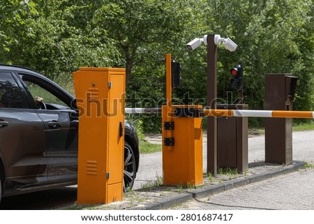 Automatic barrier gate with video camera and traffic light, as a brown car approaches the entrance in a park surrounded by trees and greenery