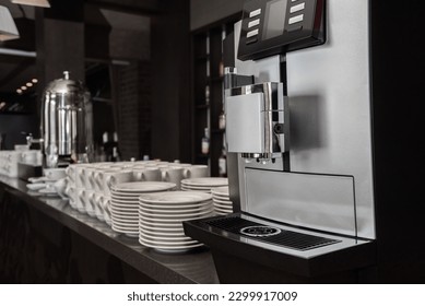 Automatic coffee machine against the background of blurred interior with mugs and plates on the bar prepared for self-service breakfast buffet.