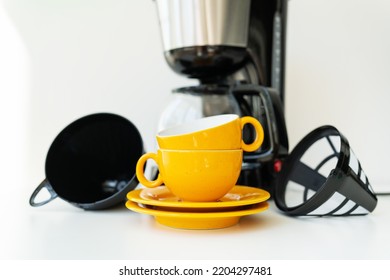 An Automatic Drip Coffee Maker Stands With A Yellow Cup On A White Table. Selective Focus, Noise. Black Modern Drip Coffee Pot. Electric Kitchen Small Household Appliances. Appliances