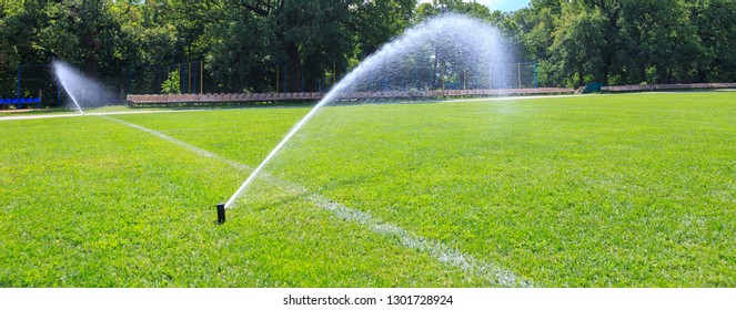 Automatic Lawn Grass Watering System At The Stadium. A Football, Soccer Field In A Small Provincial Town. Underground Sprinklers Spray Jets Water.