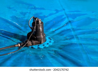 Automatic Pool Cover Pump On Top Of Blue Wet Cover. Perspective View Of Black Pool Cover Pump For Keeping The Water Away From Pool Covers Or Spas. Dirty Cover With Pine Needles. Selective Focus.