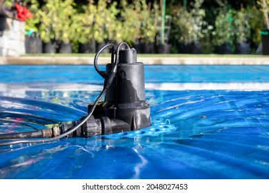 Automatic Pool Cover Pump On Top Of Blue Wet Cover In Front Of Defocused Garden Foliage. Perspective View Of Black Pool Cover Pump For Keeping The Water Away From Pool Covers Or Spas. Selective Focus.