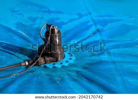 Automatic pool cover pump on top of blue wet cover. Perspective view of black pool cover pump for keeping the water away from pool covers or spas. Dirty cover with pine needles. Selective focus.