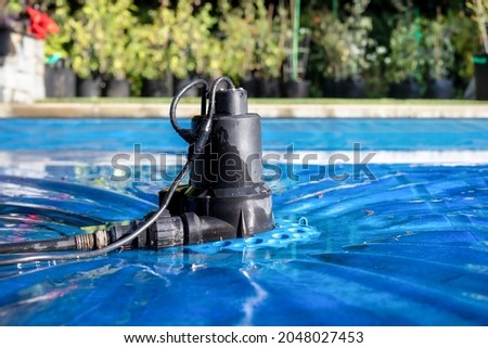Automatic pool cover pump on top of blue wet cover in front of defocused garden foliage. Perspective view of black pool cover pump for keeping the water away from pool covers or spas. Selective focus.