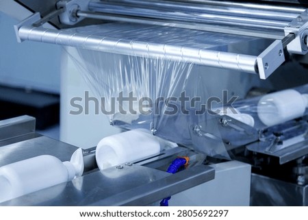 automatic shrink wrapping machine applying clear plastic film to white lotion cream pump bottles on conveyor belt in cosmetic factory. Industrial packaging, processing technology, and manufacturing.