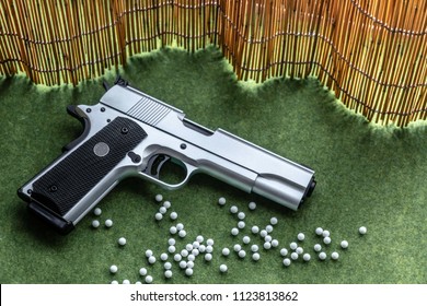 Automatic Silver Gun Airsoft guns Put on the green. There is a white BB gun on the ground. There is a bamboo backdrop.