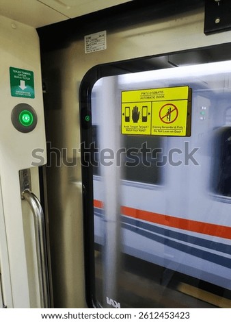 Automatic sliding door in a modern train with green-lit button and safety warning signs in Indonesian and English. Glass door reflects passing train with orange stripe design.