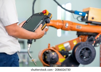 Automation Concept: Asian Senior Robot Training Engineer Is Holding Robot Control Panel And Programing Welding Robot In Workshop.