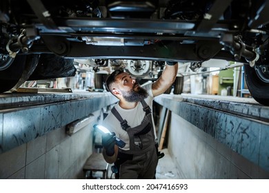 An auto-mechanic is standing in a car mechanic's pit and looking car bottom. He is lightning and looking for malfunction. A worker under the car repair workshop.