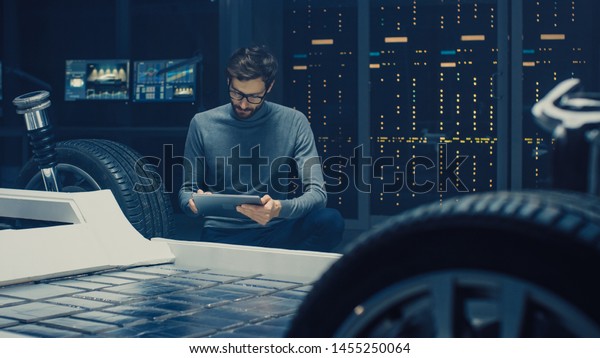 Automobile Design Engineer Sitting Beside Hybrid\
Electric Car Chassis Platform Prototype, Using Tablet Computer for\
Design Enhancement. Facility with Vehicle Frame with Suspension,\
Engine and Battery