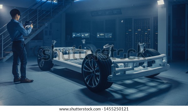 Automobile Engineer Working on Electric Car\
Chassis Platform, Using Tablet Computer Augmented Reality with 3D\
CAD Software Modelling. Innovative Facility: Vehicle Frame with\
Wheels, Engine,\
Battery