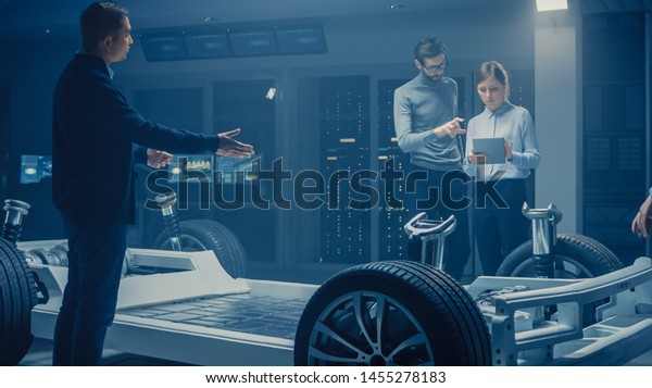 Automobile Engineers Discussing and Designing\
Electric Car Chassis Platform, Using Tablet Computers with 3D CAD\
Software. In Automotive Innovation Facility Vehicle Frame with\
Wheels Engine and\
Battery
