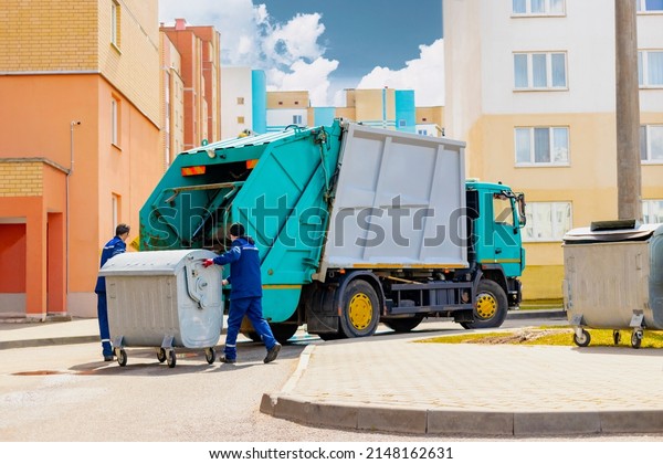 An automobile garbage\
truck collects garbage in residential areas of a modern city. Men\
load a metal container with garbage into a car for collecting and\
transporting garbage