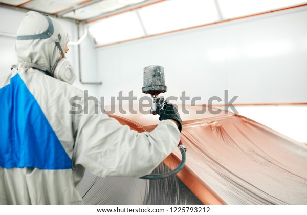 automobile painter in protective workwear\
and respirator painting car body in paint\
chamber