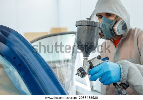 automobile repairman\
painter hand in protective glove with airbrush pulverizer painting\
car body in paint\
chamber.