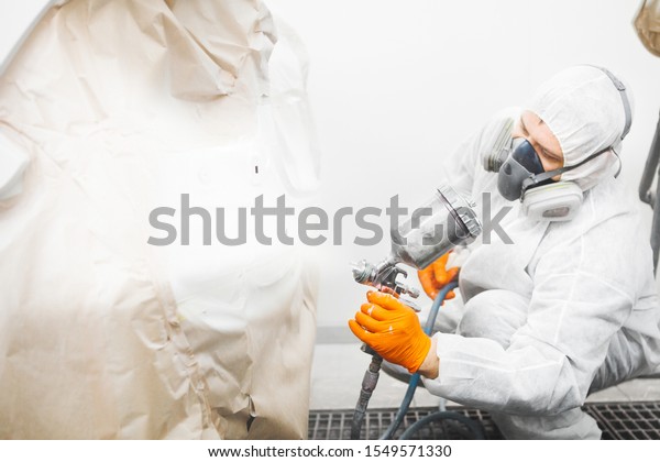 Automobile repairman painter\
in mask and protective workwear painting white car body in paint\
chamber.