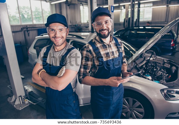 Automotive\
breakdown, wi fi internet concept. Cheerful experts at work shop,\
standing in blue safety uniform, checkered shirt, t shirt, hat head\
wear, hold pda device, background of\
vehicle