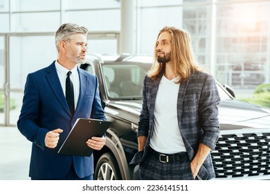 Automotive business, car sale or rental concept. Two different age men talking in a car showroom dealership with crossover on background. - Shutterstock ID 2236141511