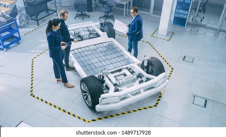Automotive Design Engineers Talking while Working on Electric Car Chassis Prototype. In Innovation Laboratory Facility Concept Vehicle Frame Includes Wheels, Suspension, Engine and Battery.