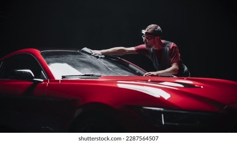 Automotive Detailer Polishing a Car Windshield with a Microfiber Towel. Specialist Wearing a Headband Lamp for Better Visual Aid for Spreading a Chemical Wax Solution Against Rain. Detailing Shop - Powered by Shutterstock