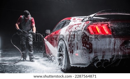 Automotive Detailer Washing Away Smart Soap and Foam with a Water High Pressure Washer. Close Up of a Red Performance Car Getting Care and Treatment at a Professional Vehicle Detailing Shop