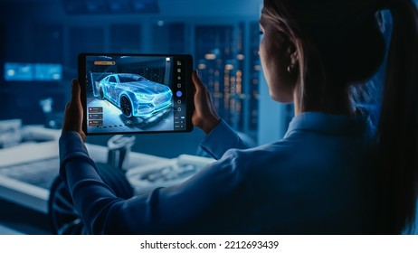 Automotive Engineer Working On Electric Car Chassis Platform, Using Tablet With Augmented Reality 3D Software. Innovative Facility: Vehicle Frame With Wheels Becomes A VFX Virtual Model.