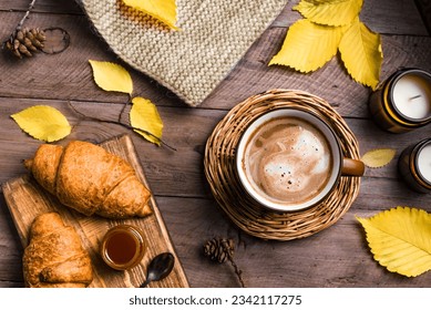 Autumn aesthetic concept. Cup of cappuccino coffee with croissants, candles, plaid and yellow autumn leaves on wooden background close up.