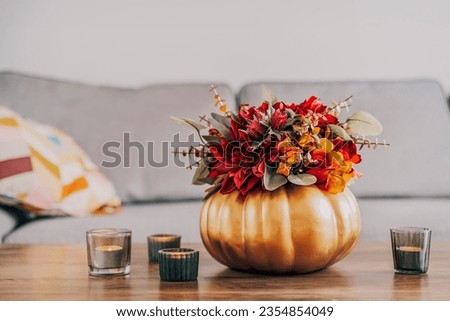 Autumn bouquet of bright artificial flowers in a golden pumpkin vase, burning candles on the wooden coffee table in living room interior with sofa. Hygge home fall decor for a cozy home atmosphere.