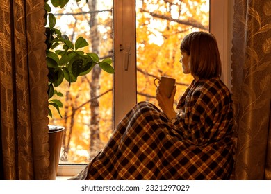 Autumn coffee. A beautiful girl with a cup of coffee in her hands sits and looks out the window. Cozy autumn composition. Copy space. - Powered by Shutterstock