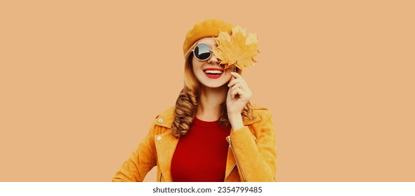 茶色のスタジオ背景にオレンジのフランスのベレー帽、ジャケットを着た黄色のカエデの葉と美しい笑顔の若い女性のポートレート、秋の色のスタイルの衣装の写真素材