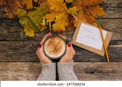 Autumn composition still life. Woman's hands with cup of coffee, cappuccino, latte coffee. autumn leaves, craft envelope and paper with eriitern Hello Autumn phrase on vintage background. Hot drink