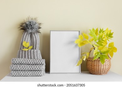 Autumn Composition With Warm Knitted Beige Scarf, Hat With Fluffy Pom Pom And Silver Blank A4 Frame With Wooden Basket And Yellow Autumn Leaves.