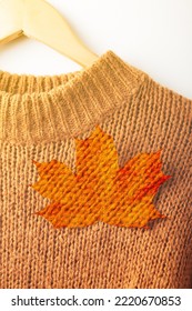 Autumn Concept. A Maple Leaf Is Attached To A Brown Chunky Knit Sweater, Close-up. The Sweater Hangs On Wooden Coat Hangers, Fragment, Close-up. Maple Leaf With Knitted Sweater Texture.