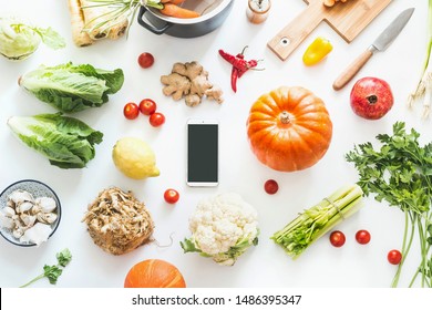Autumn cooking flat lay with smartphone with blank screen. Various seasonal fall vegetables ingredients and pumpkin on white desktop background, top view. Cozy home. Autumn harvest. Food blogging - Shutterstock ID 1486395347