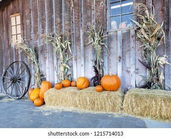An autumn decorative harvest display of pumpkins, hay bails, corn stalks, and a wagon wheel against a wooden barn. 
