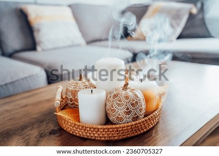 Autumn fall cozy mood composition for hygge home decor. Orange pumpkins decorated with mandalas, white blown candles with smoke on wicker tray on the coffee table in the living room. Selective focus.