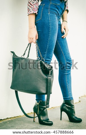 Autumn fashion outfit. Fashionable woman long legs in denim pants black stylish high heels shoes and handbag outdoor on city street