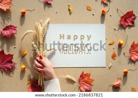 Autumn frame, caption greeting text Happy Holidays. Natural Fall leaves, wheat ears in hand with manicure nails. Natural terracotta, beige, yellow and red color shades. Flat lay. top view.