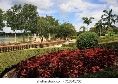 Autumn garden featuring red leafed shrubs among green hedges and trees, with a stone wall and lakefront patio, and palm trees.