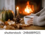 Autumn hygge. Hands holding cup of tea and pumpkin, autumn leaves on table on background of warm fireplace. Cozy fall in living room. Thanksgiving and Halloween.