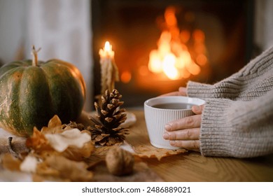 Autumn hygge. Hands holding cup of tea and pumpkin, autumn leaves on table on background of warm fireplace. Cozy fall in living room. Thanksgiving and Halloween.