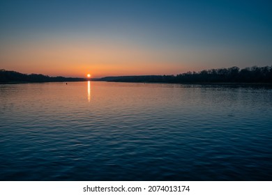 An Autumn Landscape At Sunset On A Wide River With Bright Red Reflections And A Sun Disc Over The Horizon. The Rural Scene Photo From A Low Point View.