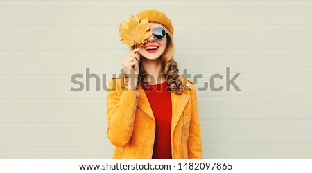 Autumn mood! happy smiling woman holding in her hands yellow maple leaves covering her eye over gray wall background